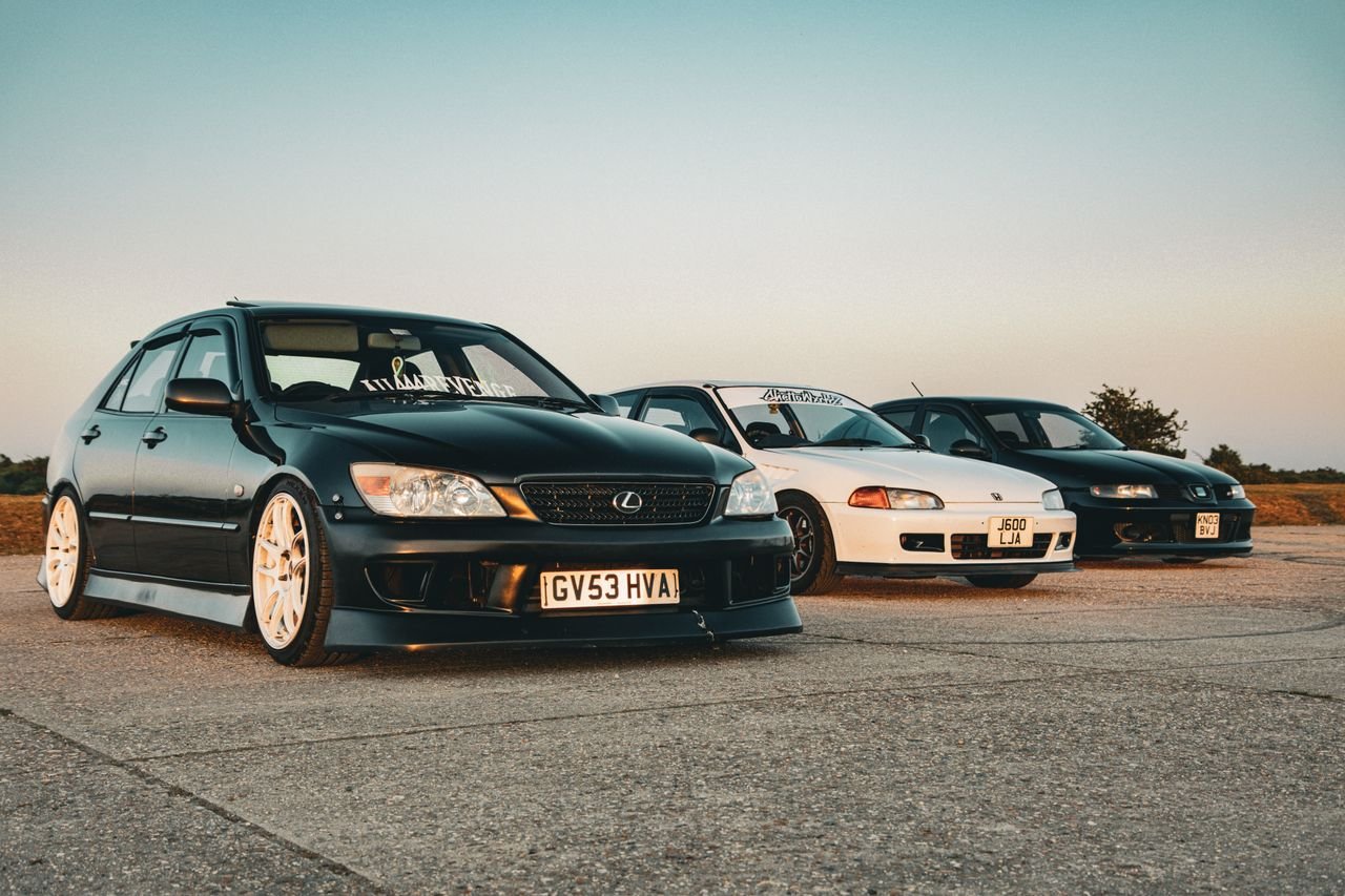 A line-up of custom sports cars including Lexus and Honda models on a road at sunset.