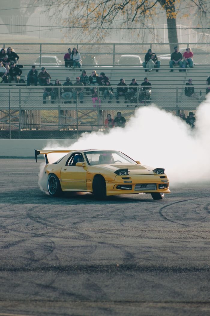 Yellow Mazda RX-7 drifts on track with spectators watching from stands.